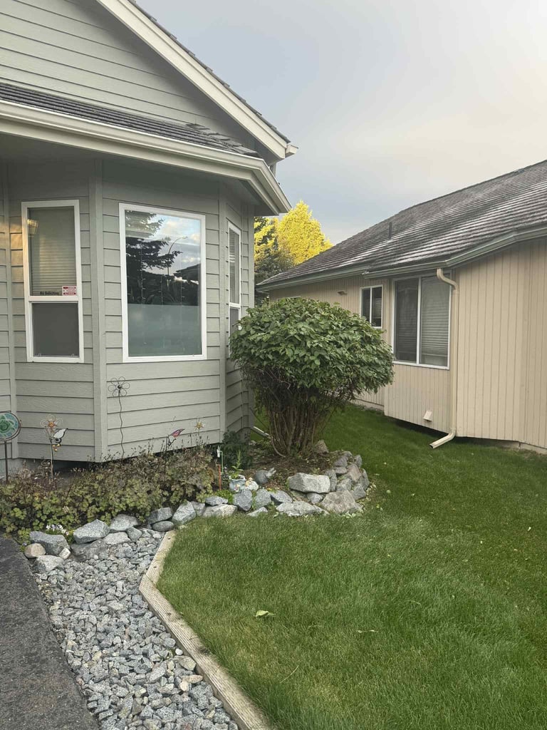 Modern house exterior with gray siding, white-trimmed windows, landscaped yard with green lawn, gravel pathways, and shrubs